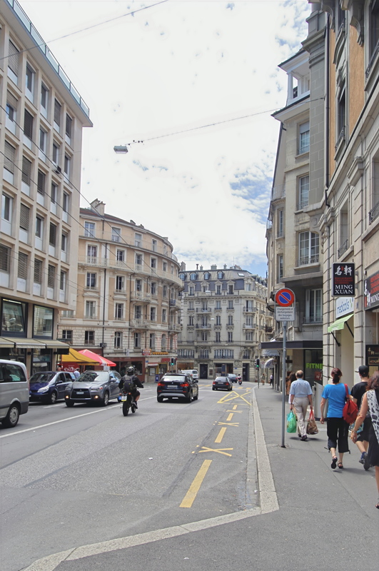4 - A sloping urban street scene in Lausanne, Switzerland, featuring European-style multi-story apartment buildings, passing traffic, and pedestrians